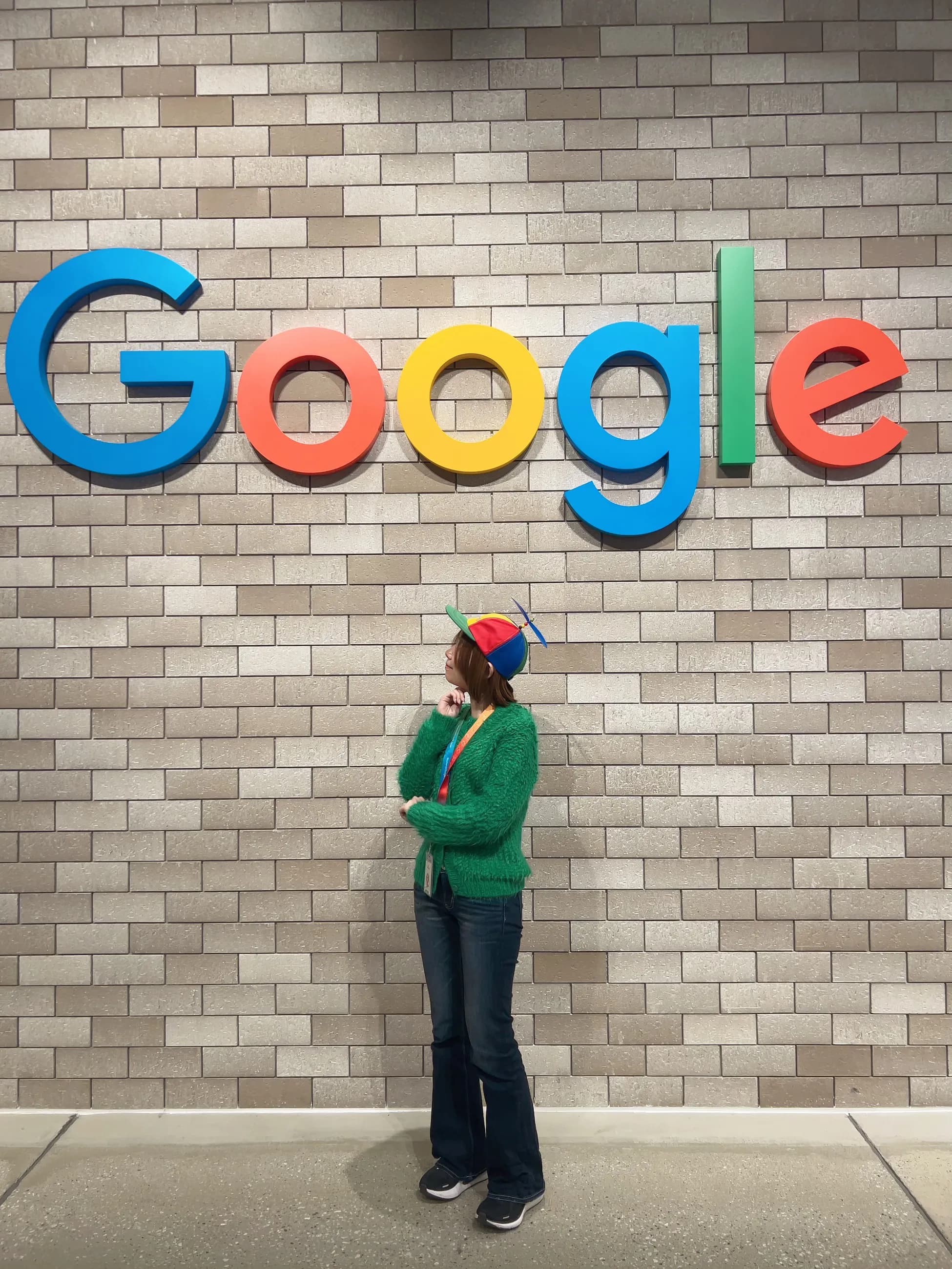 Google オフィスのロゴの前で、プロペラ付きの帽子をかぶって立つ様子